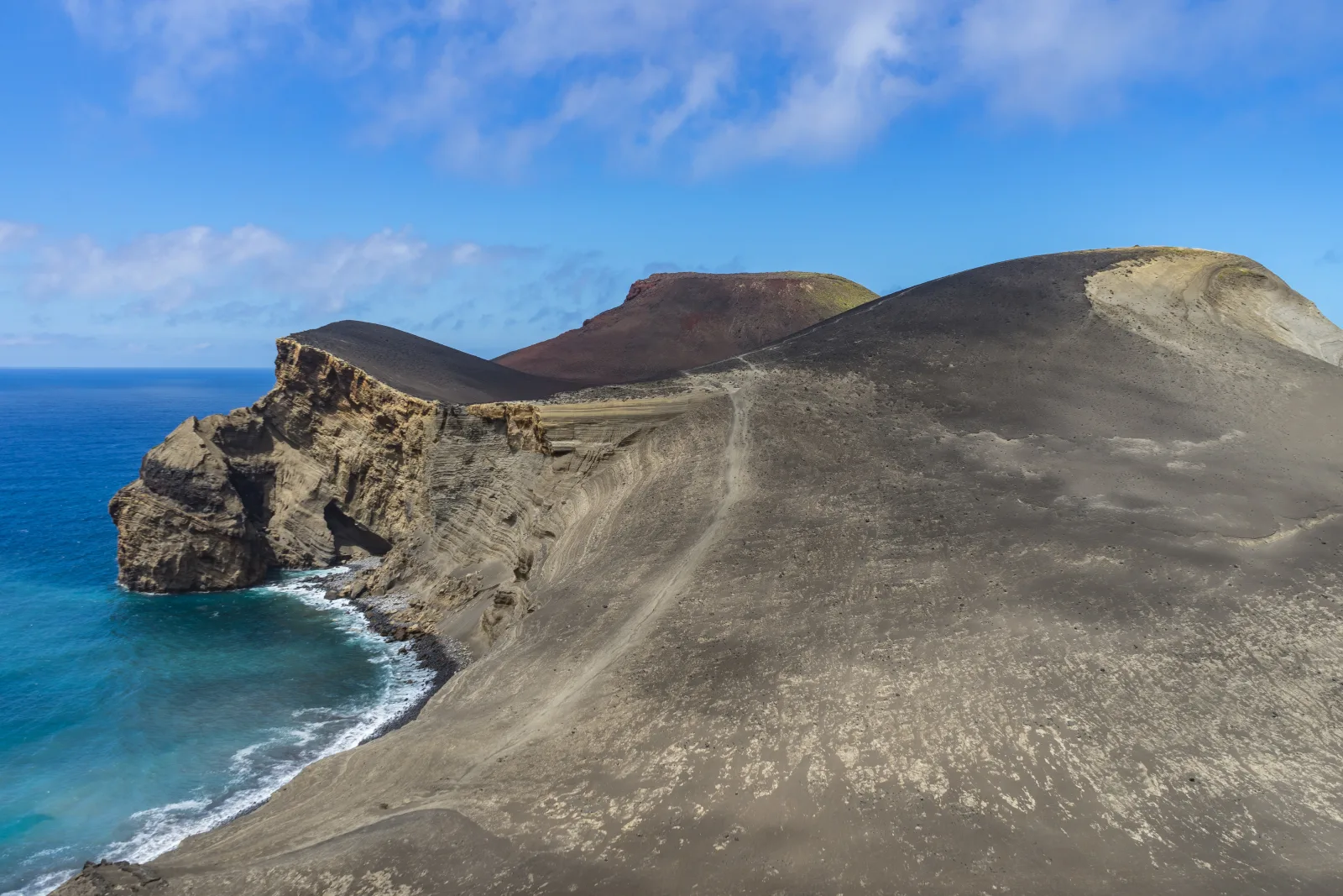 The arid moonscape of Capelinhos — black volcanic ash deposits left by the 1957–58 eruption, with the half-buried Farol dos Capelinhos lighthouse standing in the distance, Faial Island, Azores